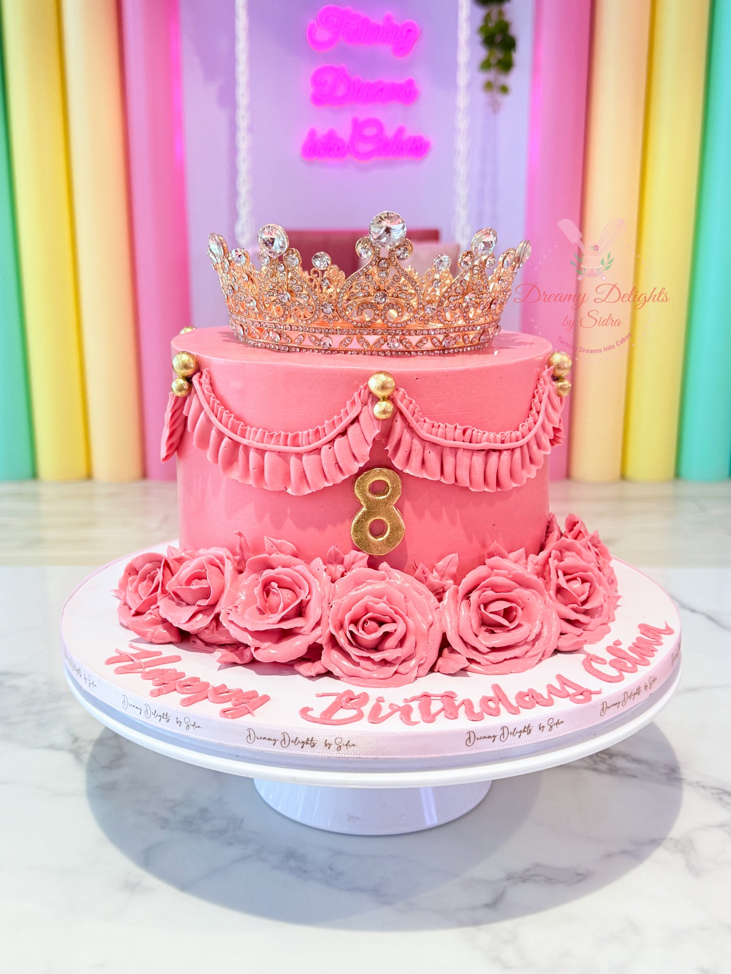 Pink birthday cake with a tiara on top, decorated with roses and the number 8, against a colorful background.