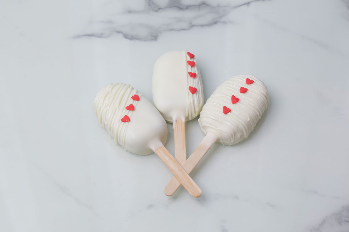 Mini heart-shaped Valentine’s Day cakesicles with white coating and red heart details by Dreamy Delight by Sidra