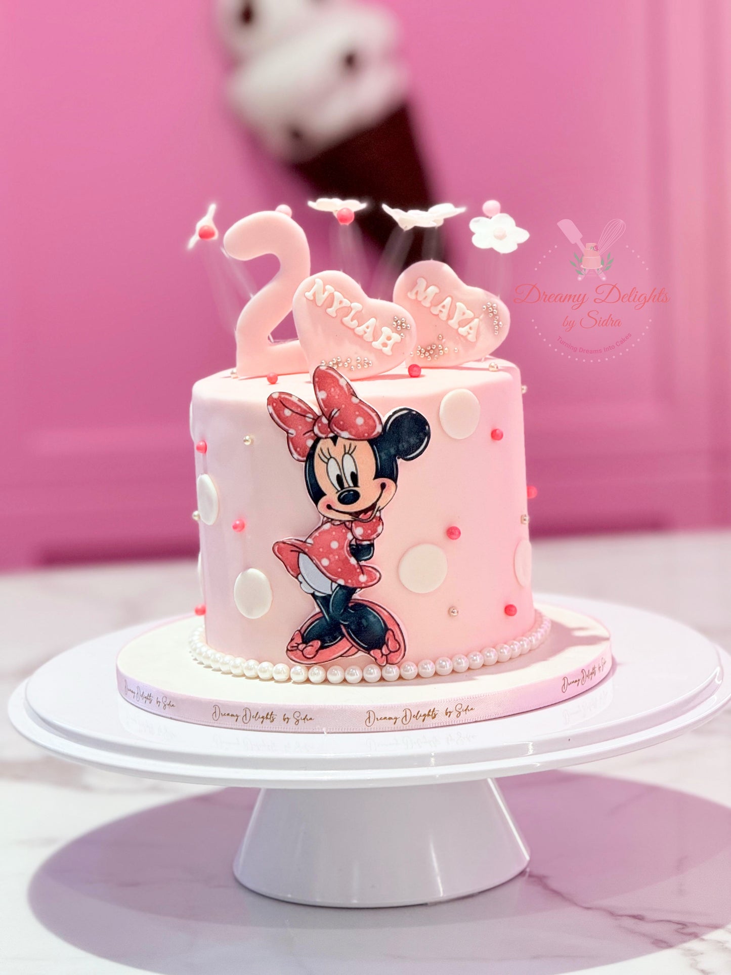 Pink birthday cake with Minnie Mouse design on a white stand against a pink background