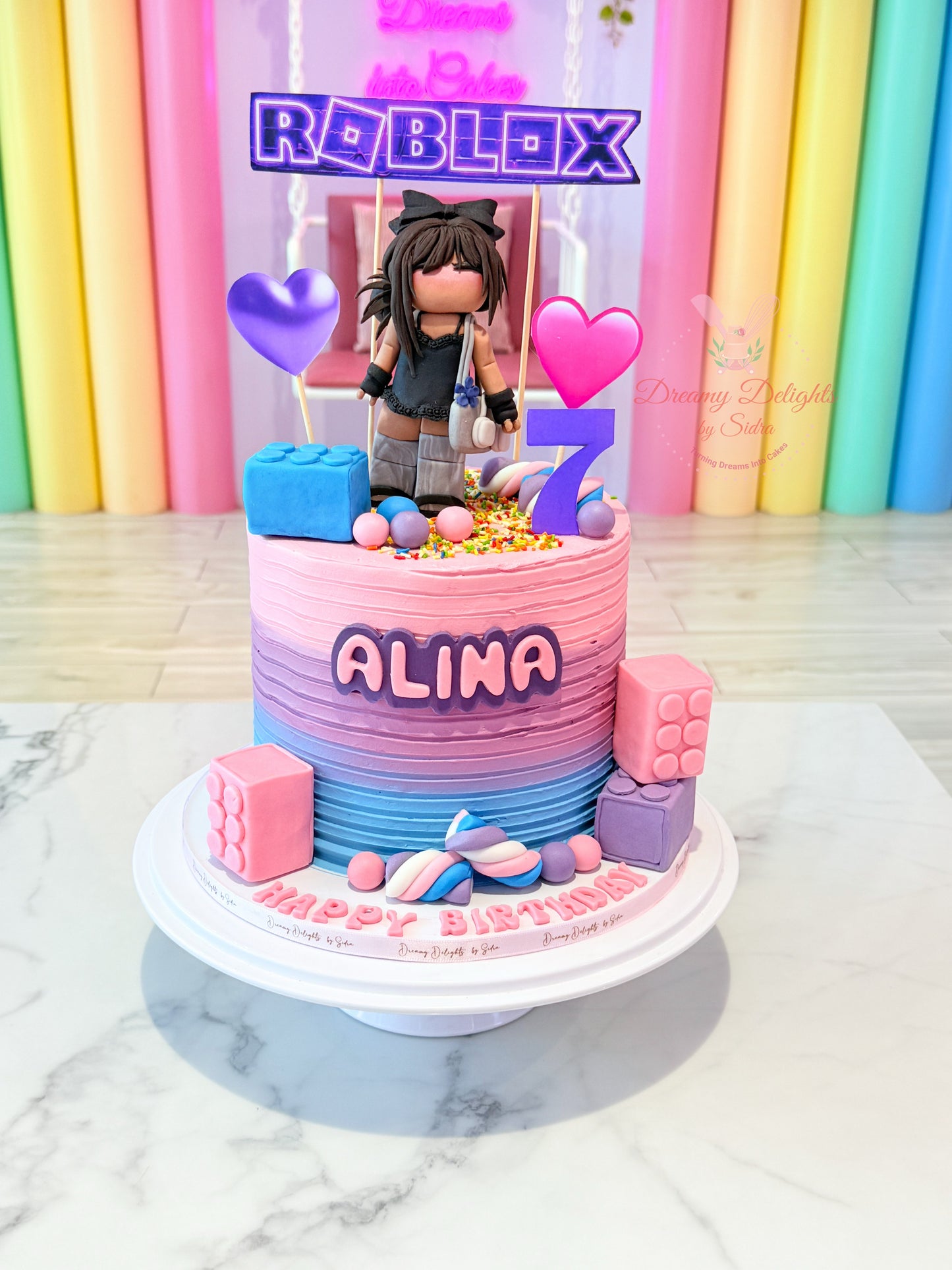 Colorful birthday cake with Roblox character and 'Alina' on a marble surface with rainbow curtains in the background.