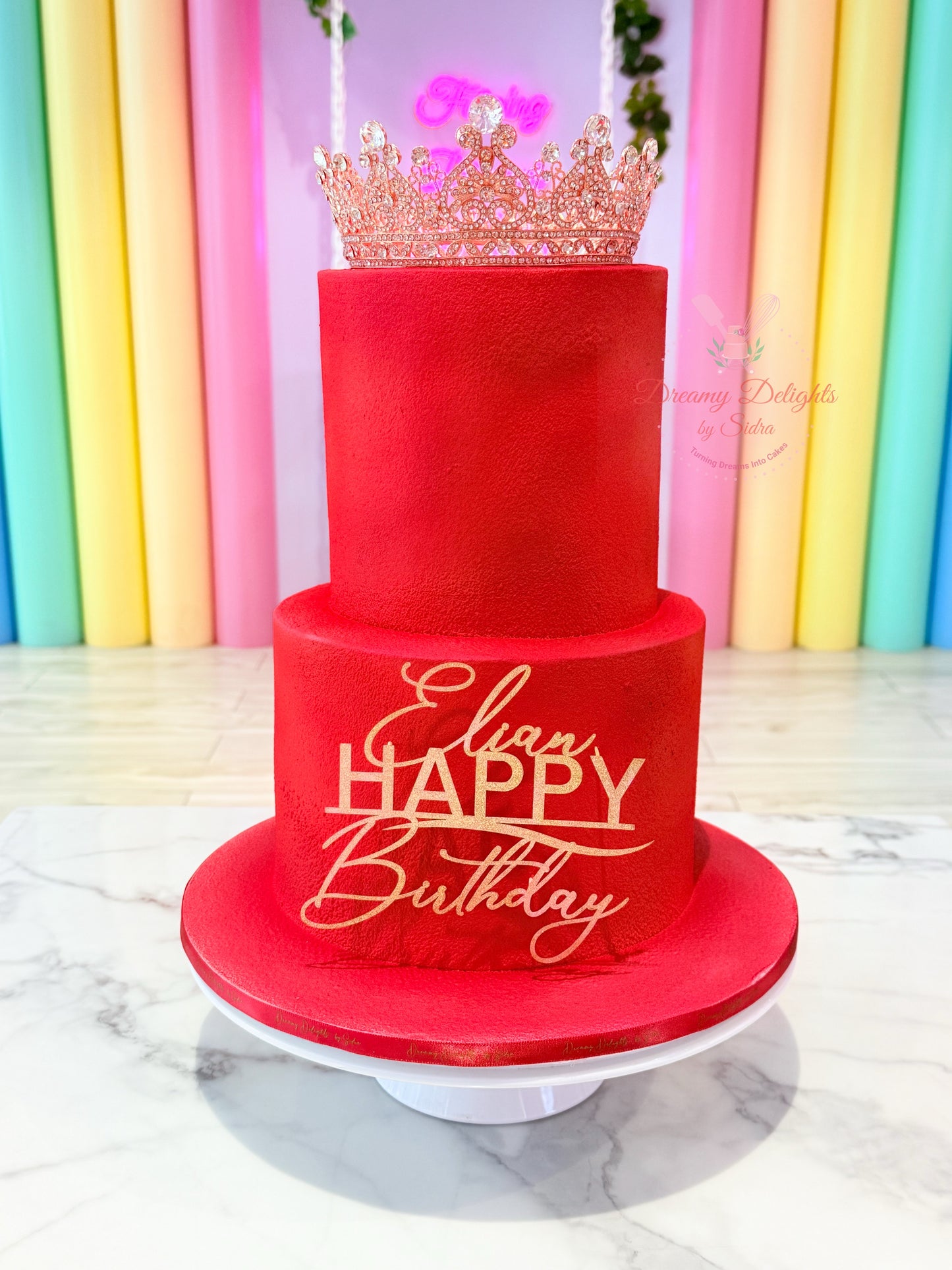 Red birthday cake with 'Happy Birthday' and a tiara on top, against a colorful striped background.