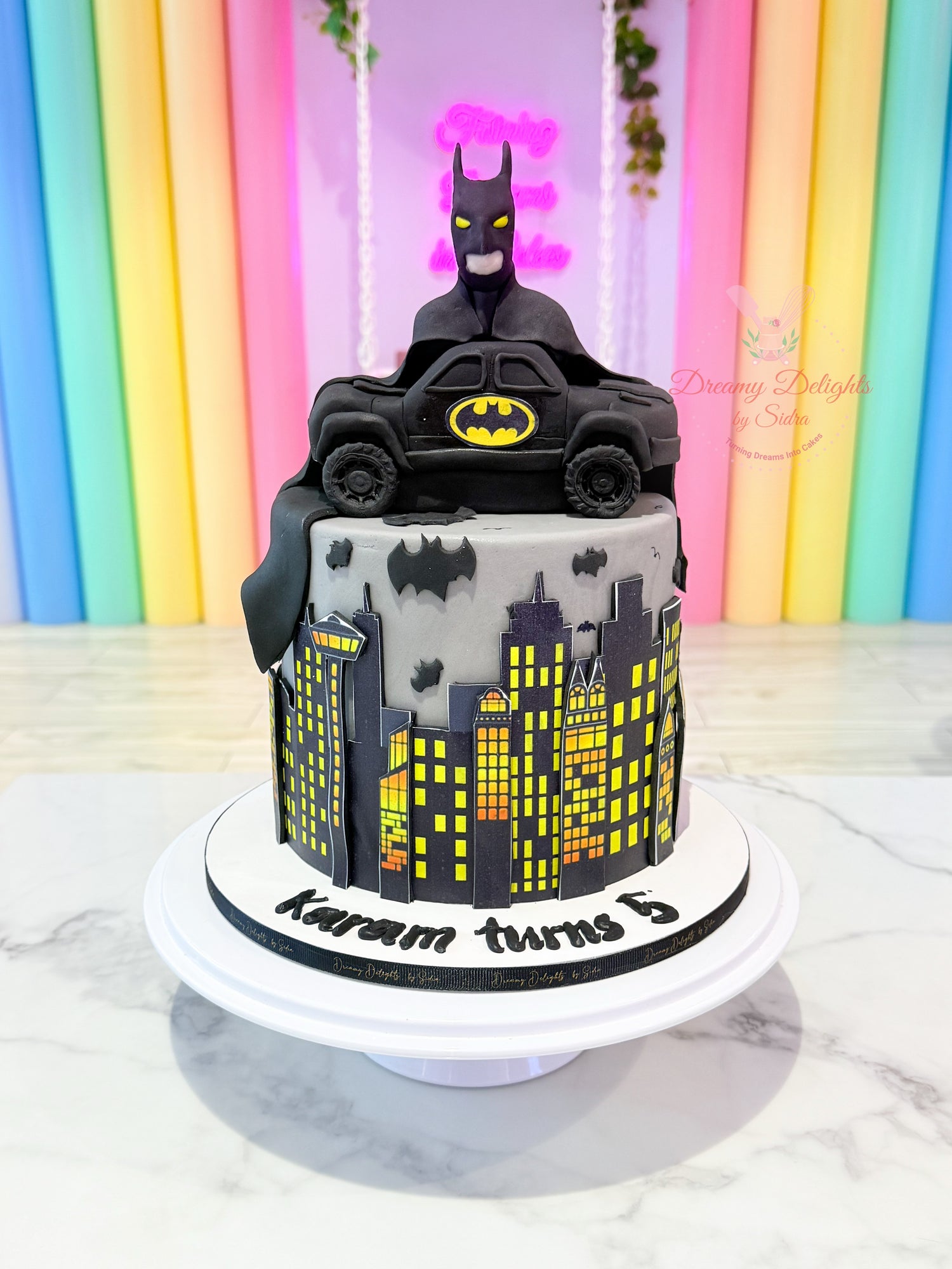 Batman-themed birthday cake with a toy car and cityscape design against a colorful background.