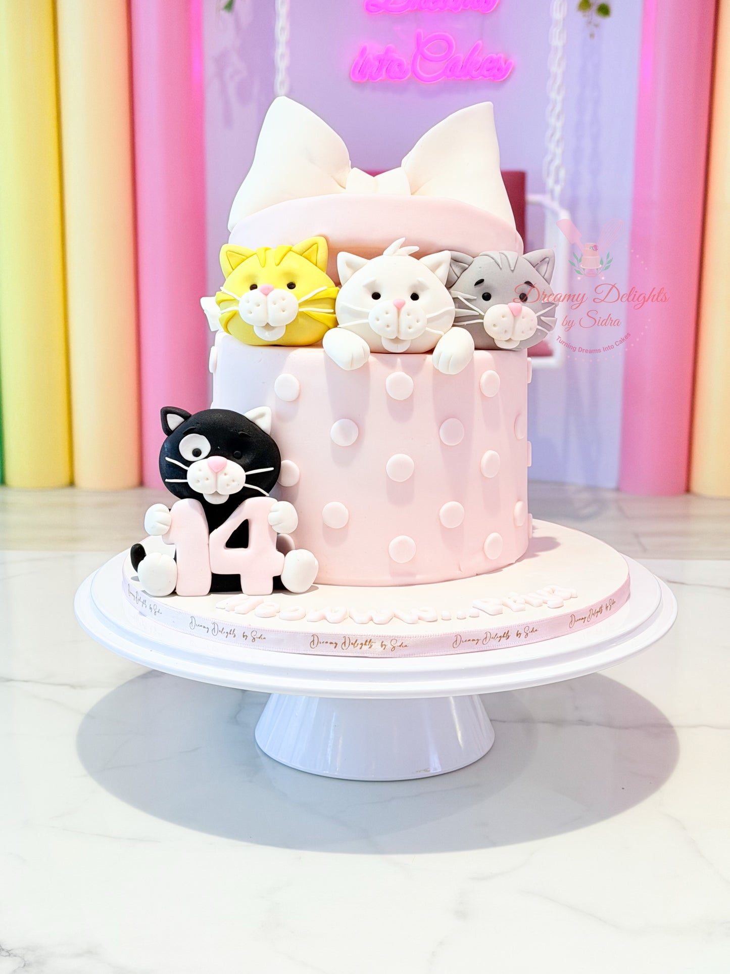 Pink cake with cat-themed decorations on a white stand against a colorful background