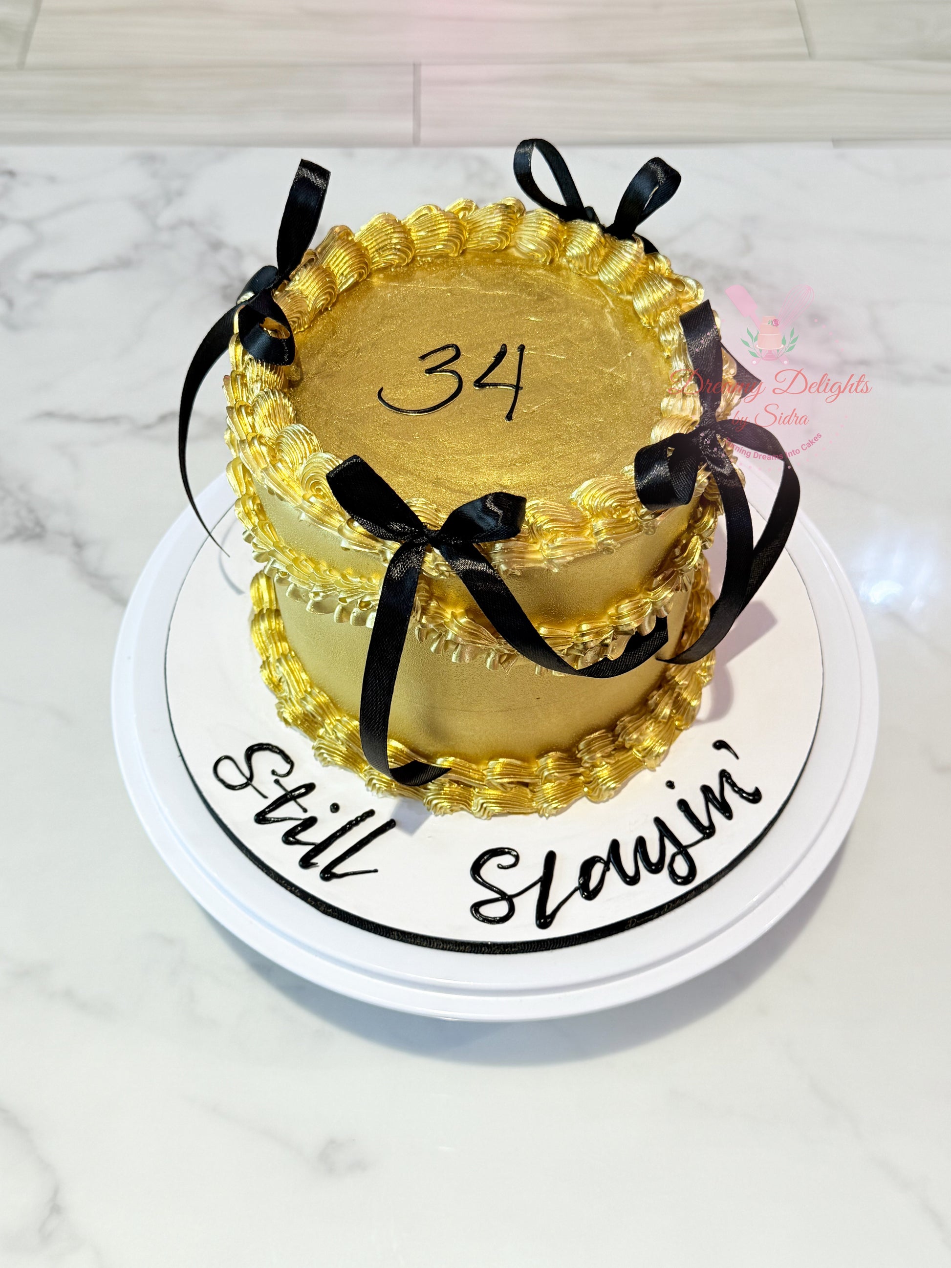 Gold birthday cake with number 34 and 'Still Slayin'' text on a white marble background