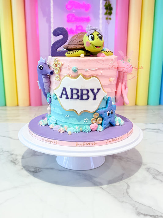 Birthday cake with sea-themed decorations on a stand, against a colorful background.