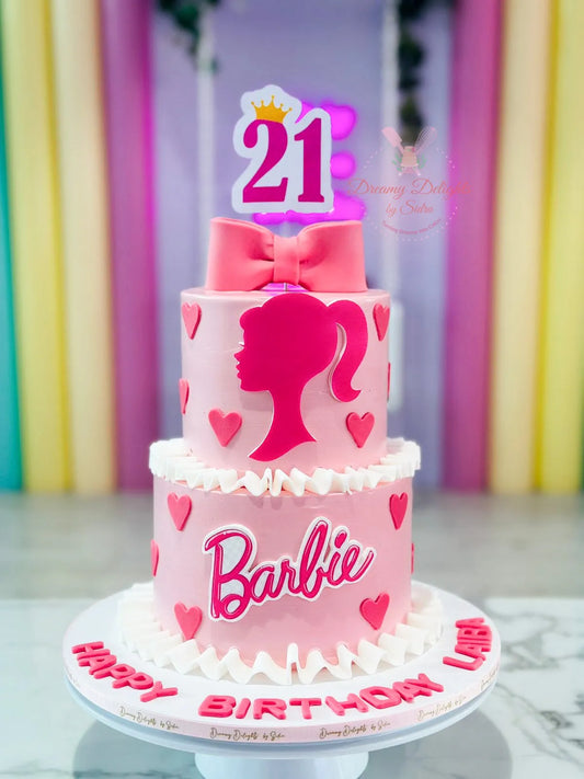 A two-tier Barbie-themed cake decorated with pink and white fondant, featuring the Barbie logo and a number '21' on the top tier, with a 'Happy Birthday' sign on the bottom tier.