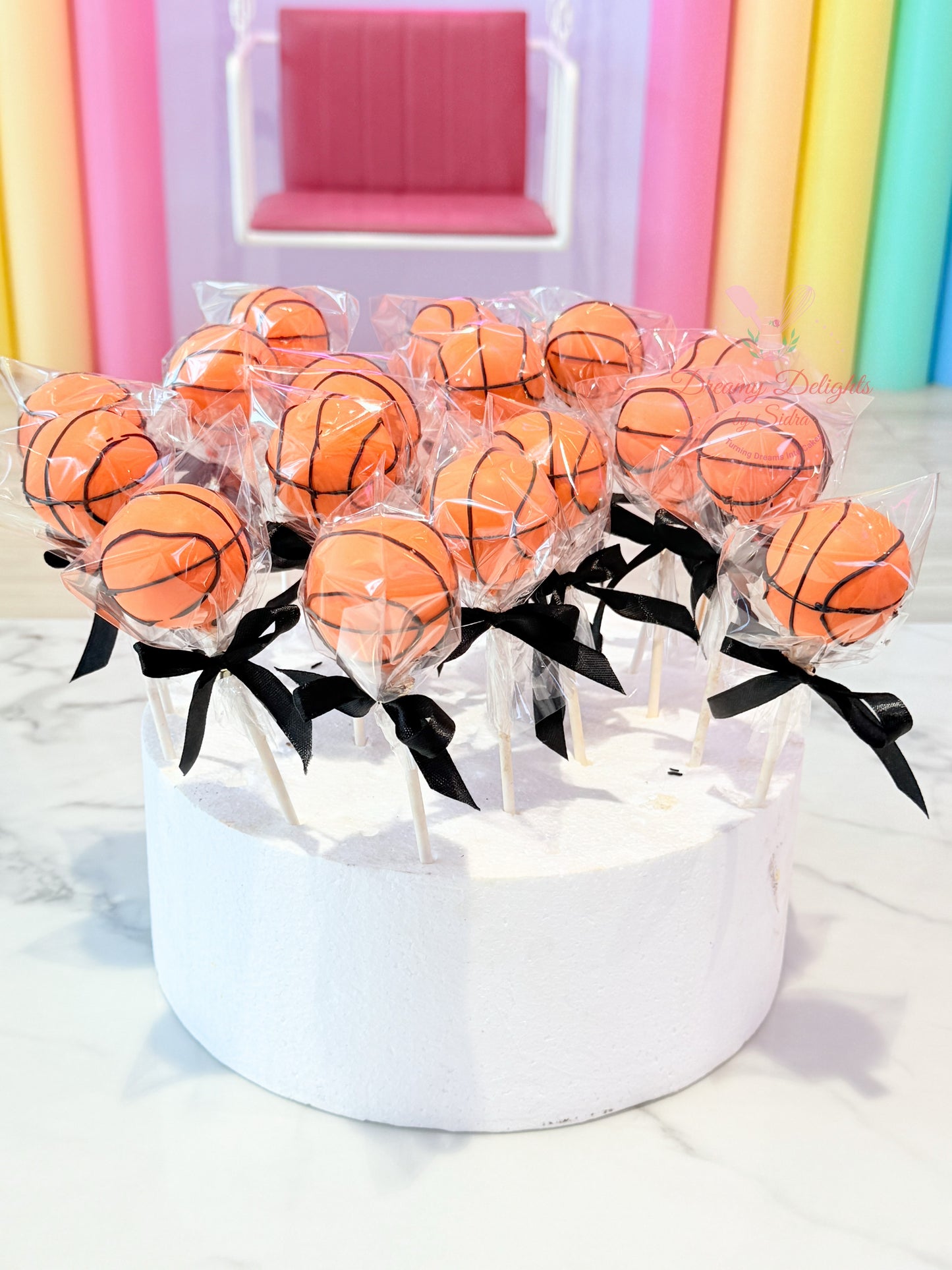 Basketball-shaped lollipops on sticks with black ribbons in a white box against a colorful curtain background.