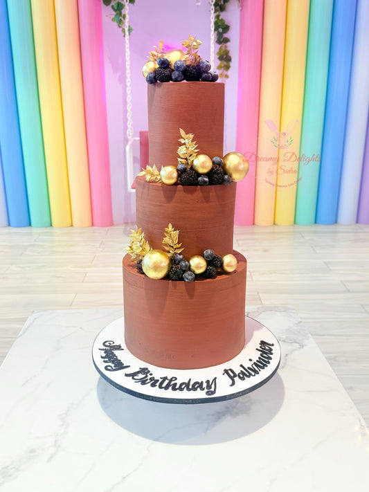 Three-tiered chocolate cake with gold and black decorations against a colorful curtain background