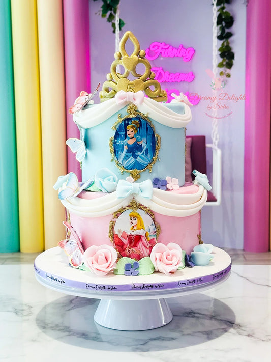 A decorated cake with character figures of Princess Cinderella and Princess Aurora, presented on a display stand, surrounded by floral decorations.