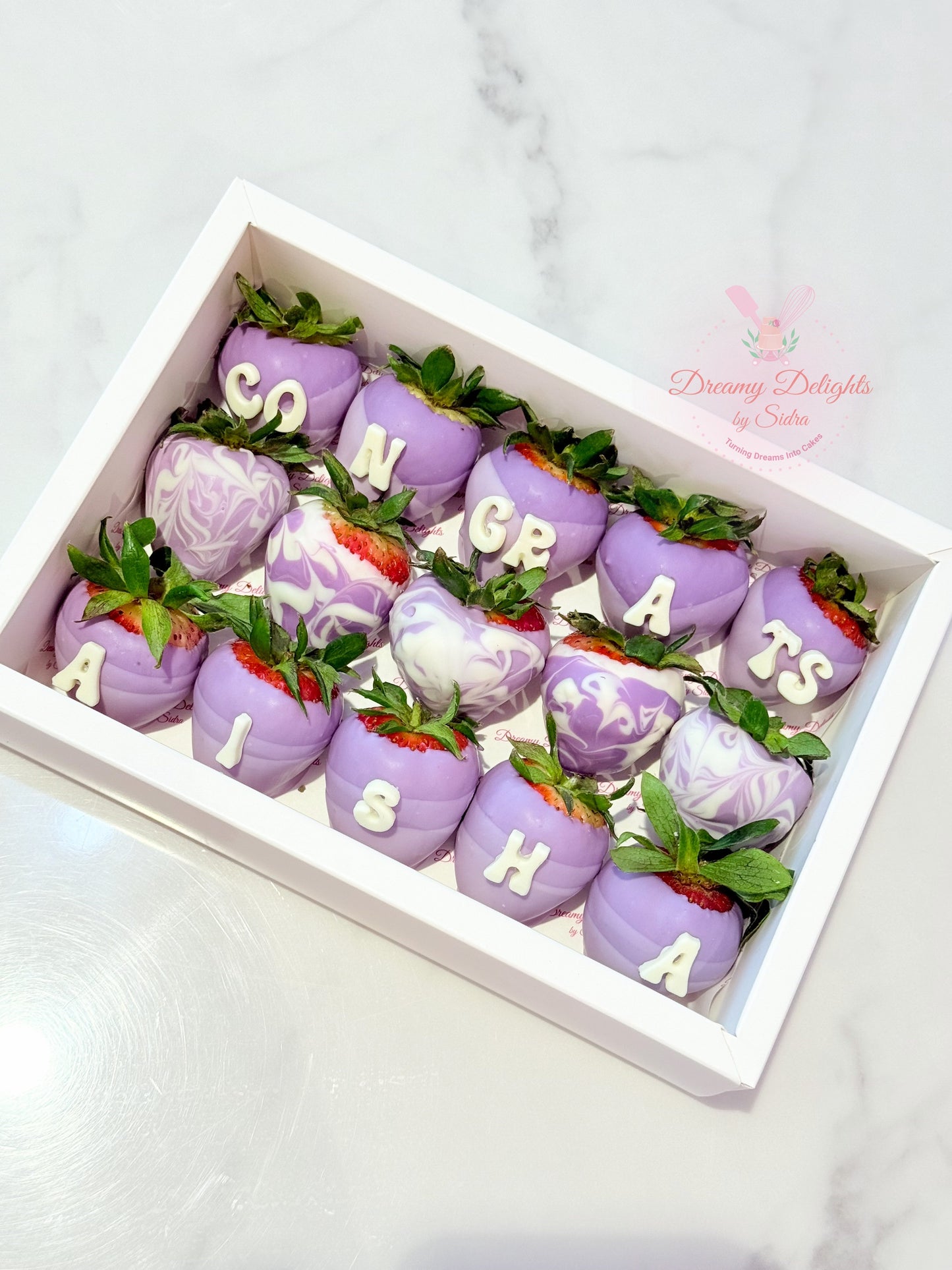 Decorative strawberries with purple icing and letter decorations in a box, on a light gray background.