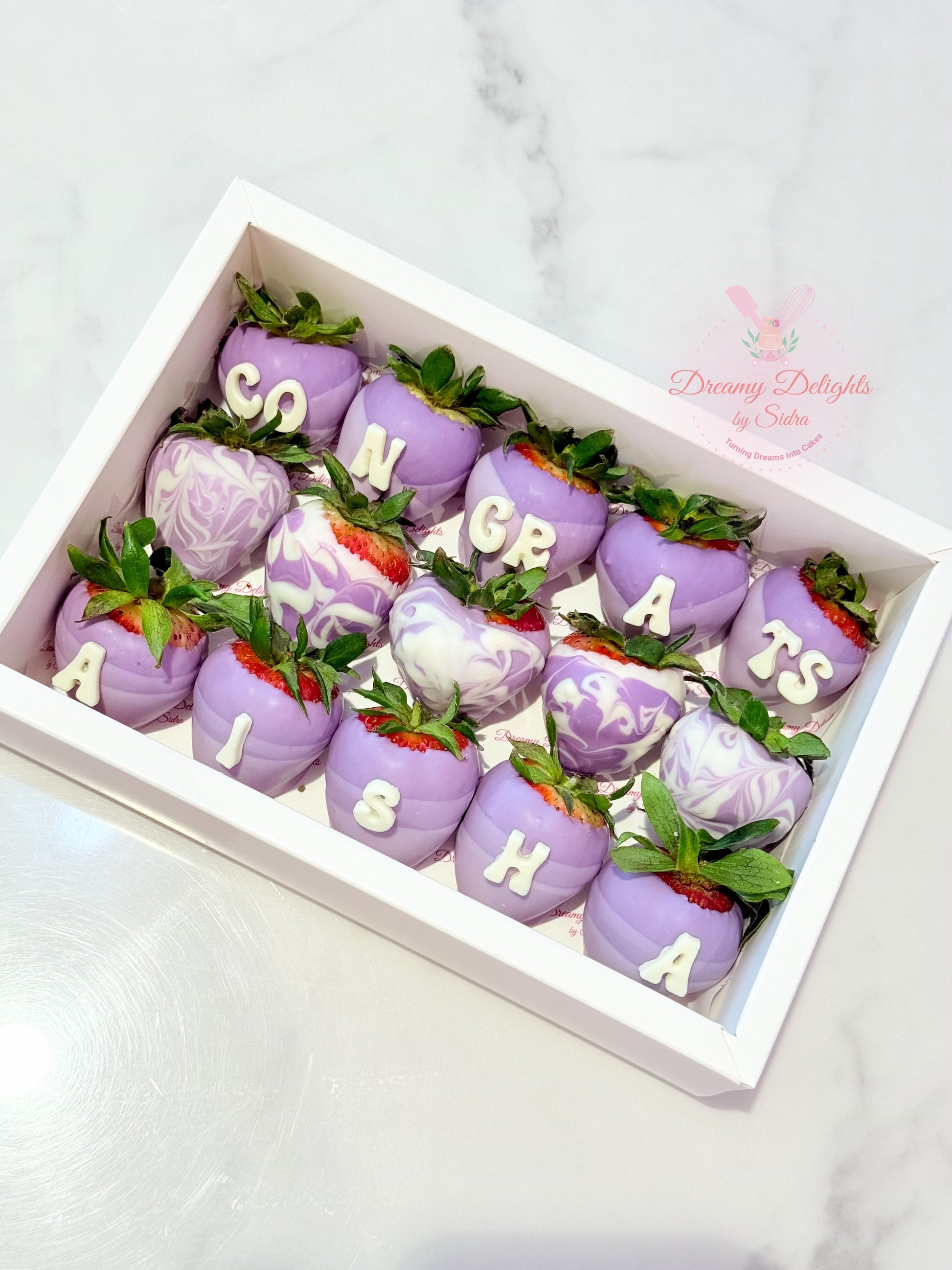 Decorative strawberries with purple icing and letter decorations in a box, on a light gray background.