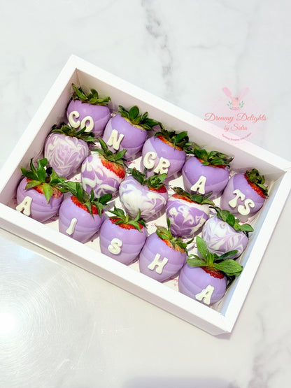 Decorative strawberries with purple icing and letter decorations in a box, on a light gray background.