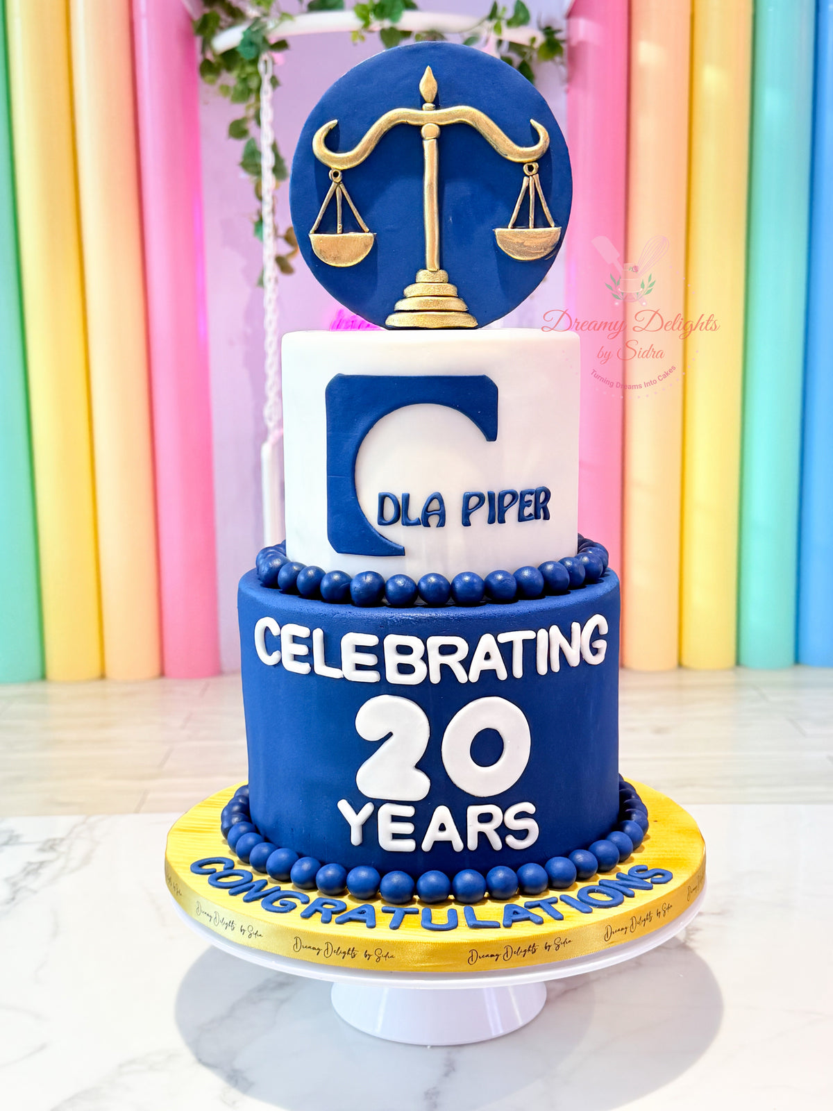 Corporate anniversary cake Dubai with blue tiers, gold scales and company branding by Dreamy Delights by Sidra.