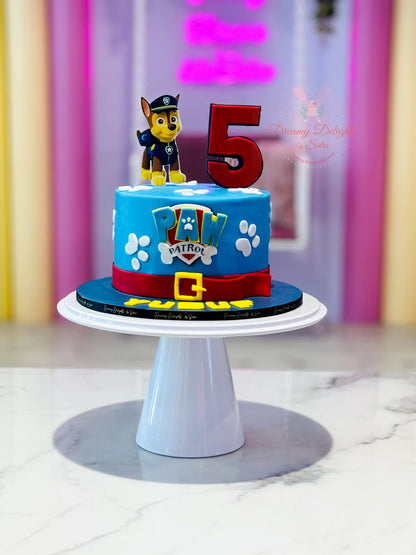 A Paw Patrol themed birthday cake with a blue and red design, featuring a Paw Patrol character on top, placed on a white stand.