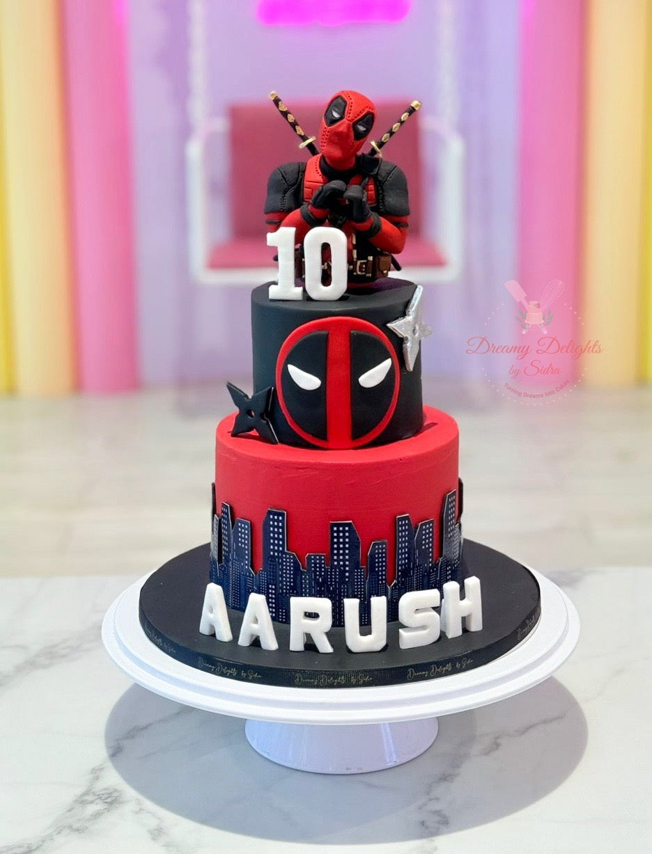 Two-tier birthday cake with Deadpool figure and 'Aarush' on a marble surface.