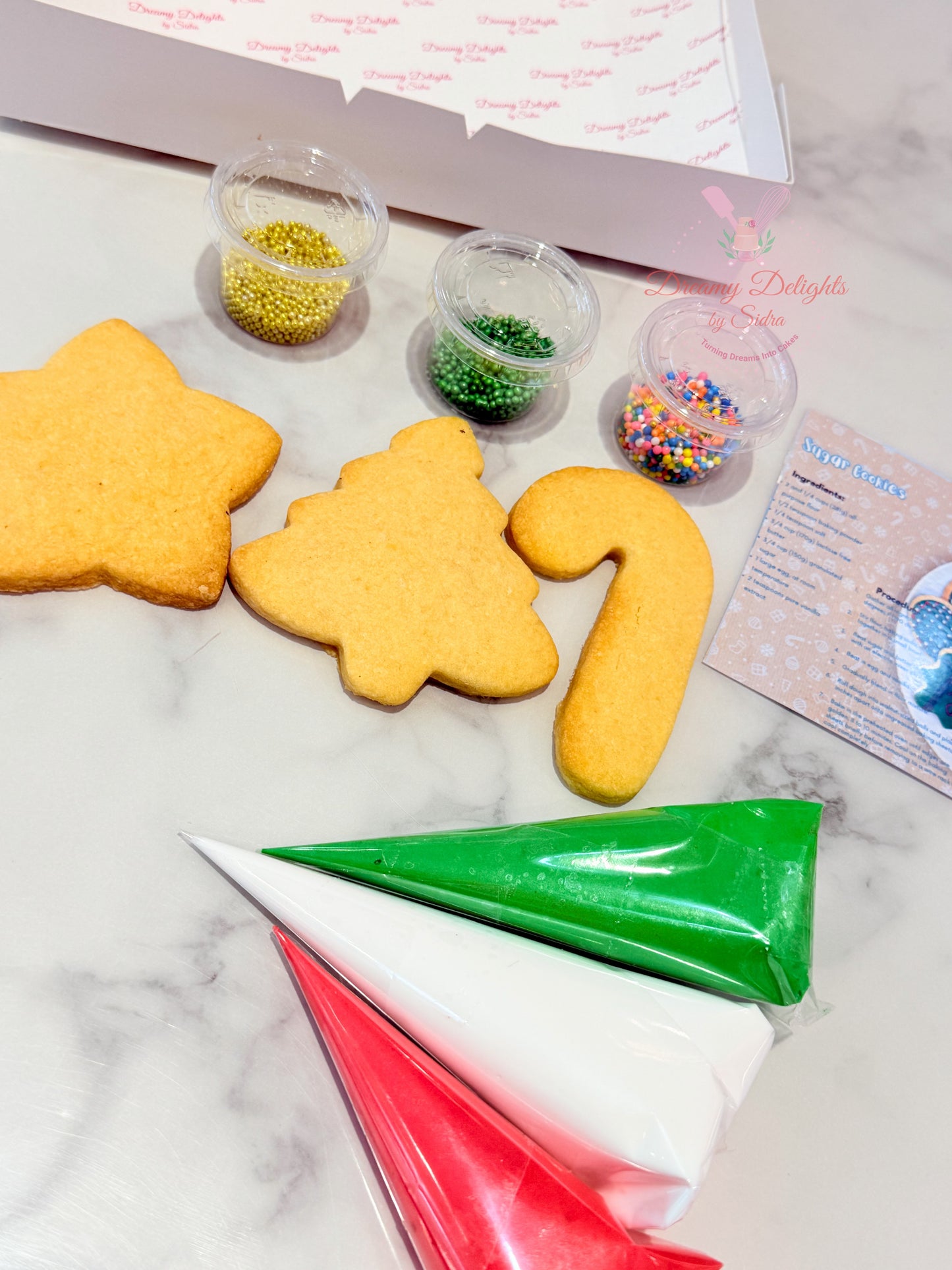 Decorative cookies shaped like a star, tree, and number '12' with icing bags and sprinkles on a marble surface.