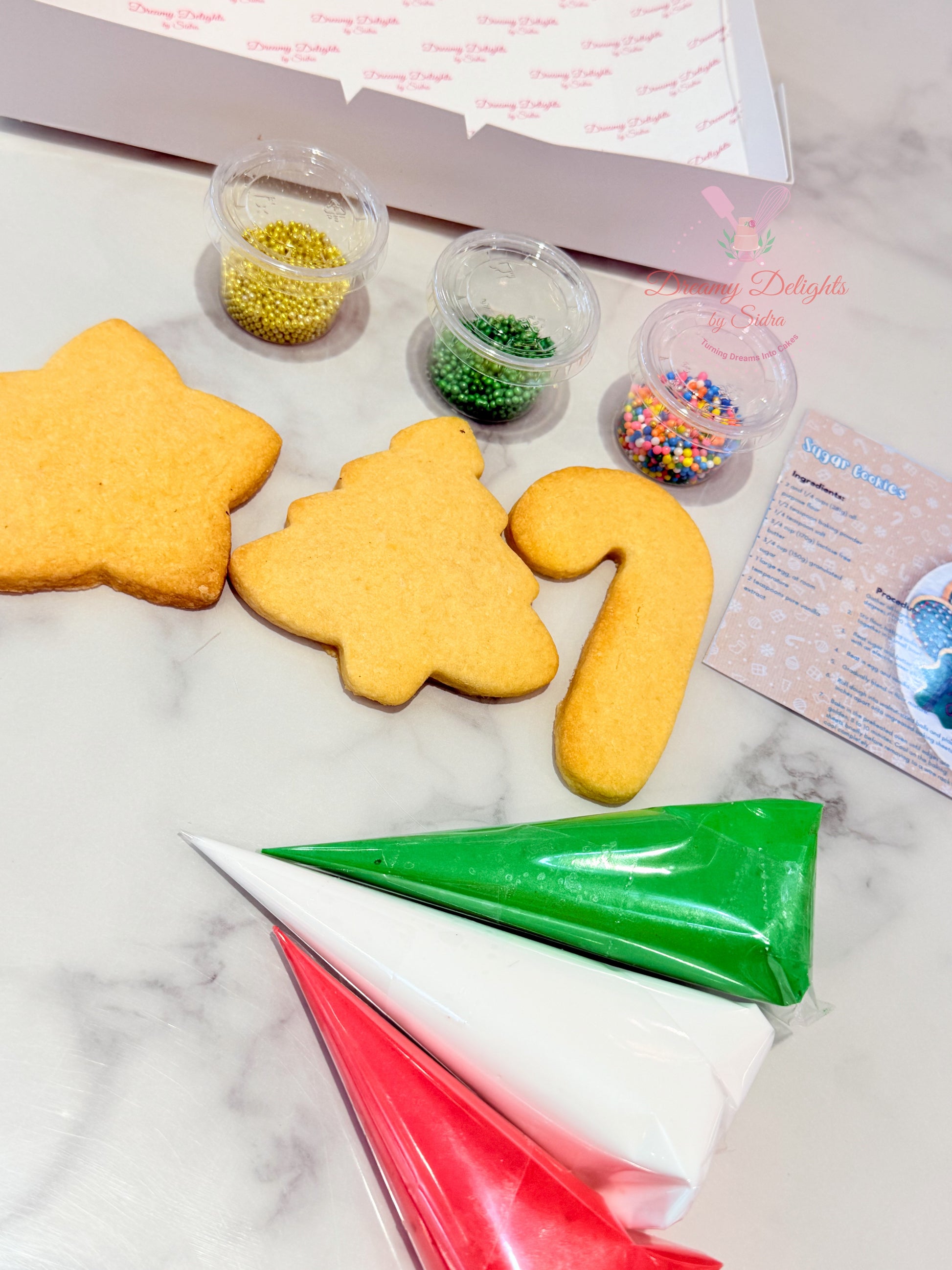 Decorative cookies shaped like a star, tree, and number '12' with icing bags and sprinkles on a marble surface.