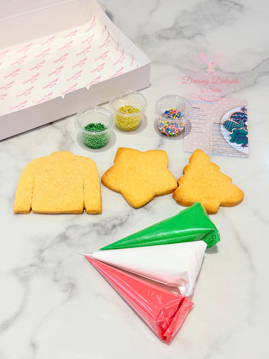 Decorative cookies shaped like a sweater, star, and tree with icing bags and sprinkles on a marble surface.