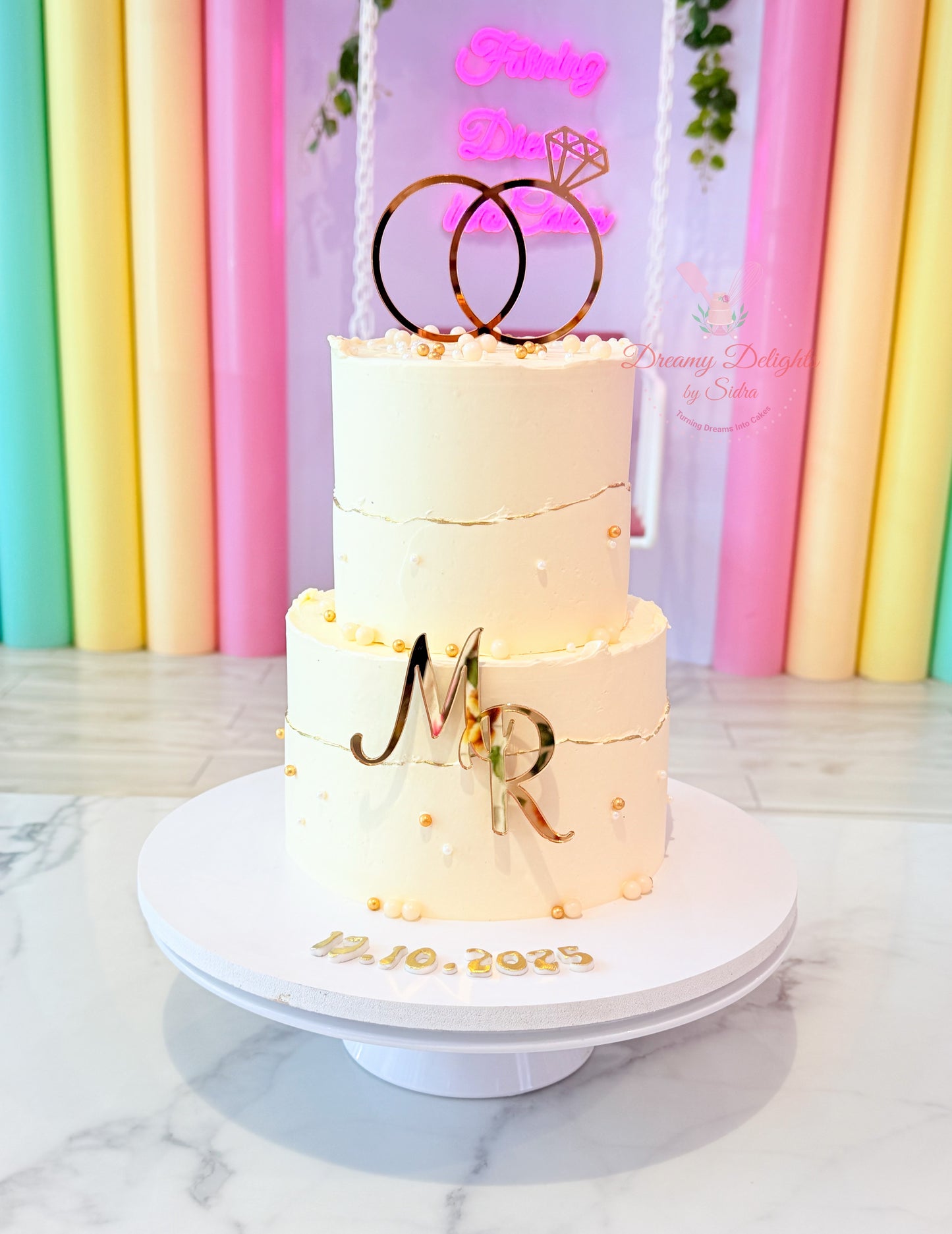 Elegant two-tier engagement cake with gold ring topper, personalized initials, white buttercream finish, and gold pearl decorations in Dubai.