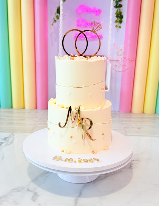 Elegant two-tier engagement cake with gold ring topper, personalized initials, white buttercream finish, and gold pearl decorations in Dubai.