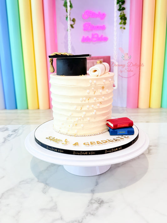Elegant graduation degree cake with white ruffled frosting, gold accents, graduation cap and diploma decorations by Dreamy Delights by Sidra in Dubai.