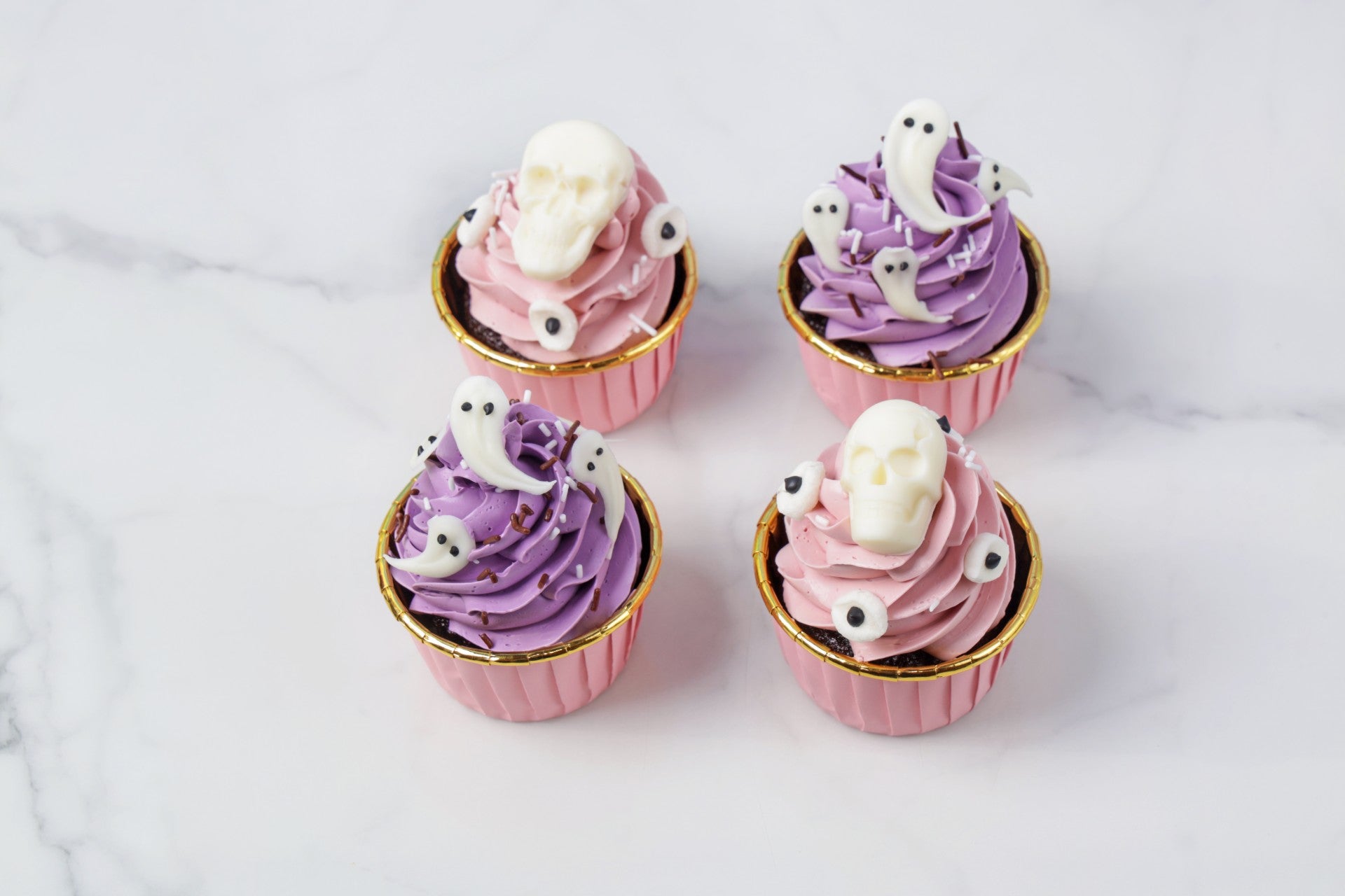 Four cupcakes with purple and pink frosting and ghost decorations on a white background
