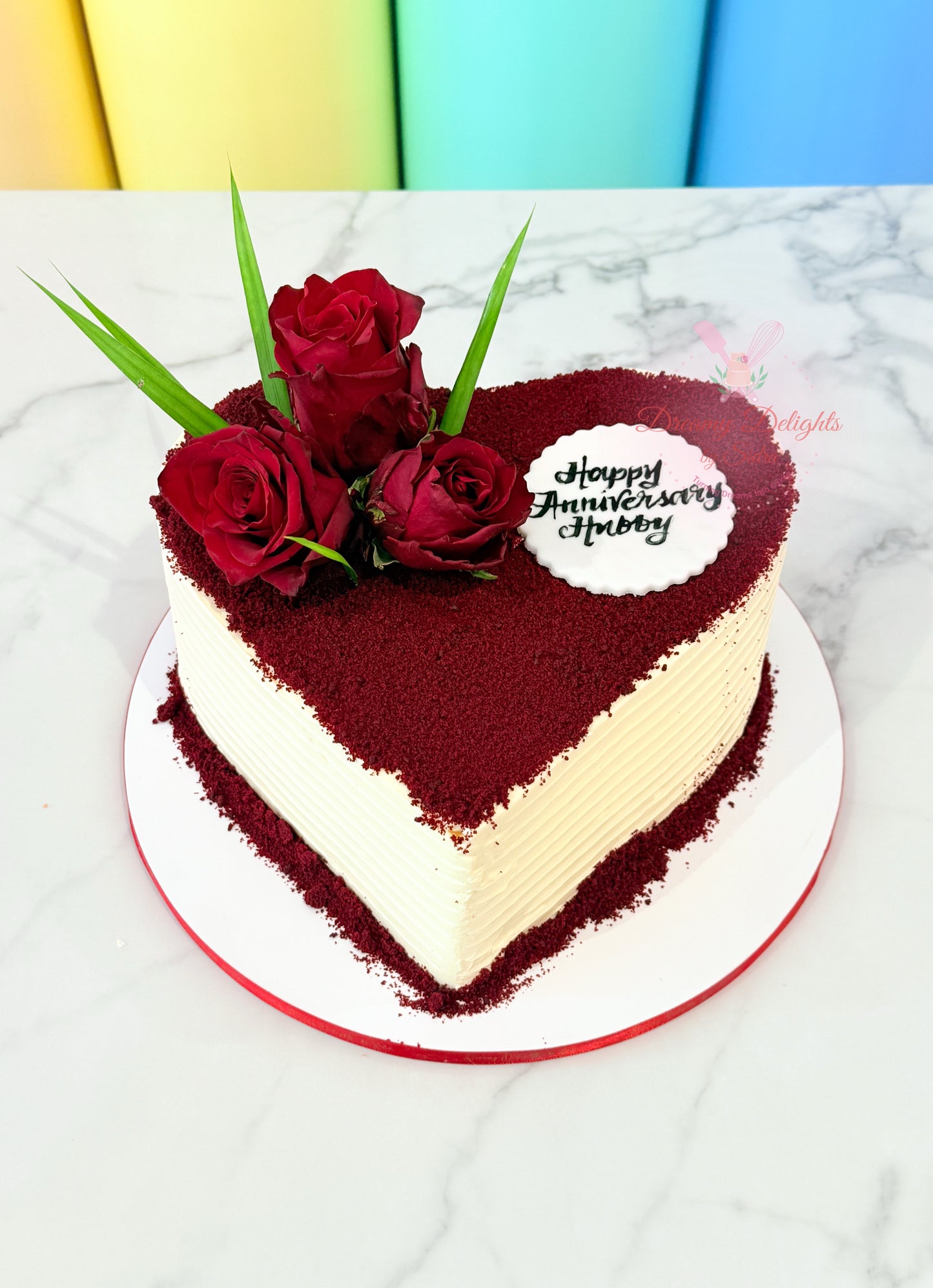 Heart-shaped cake with red roses and a 'Happy Anniversary Hubby' message on a marble surface.