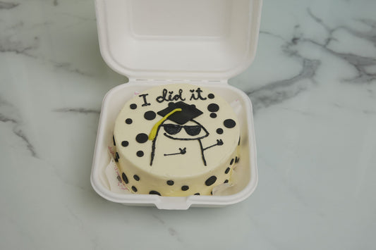 Small round cake with polka dots and a graduation-themed design in a white container on a marble surface.