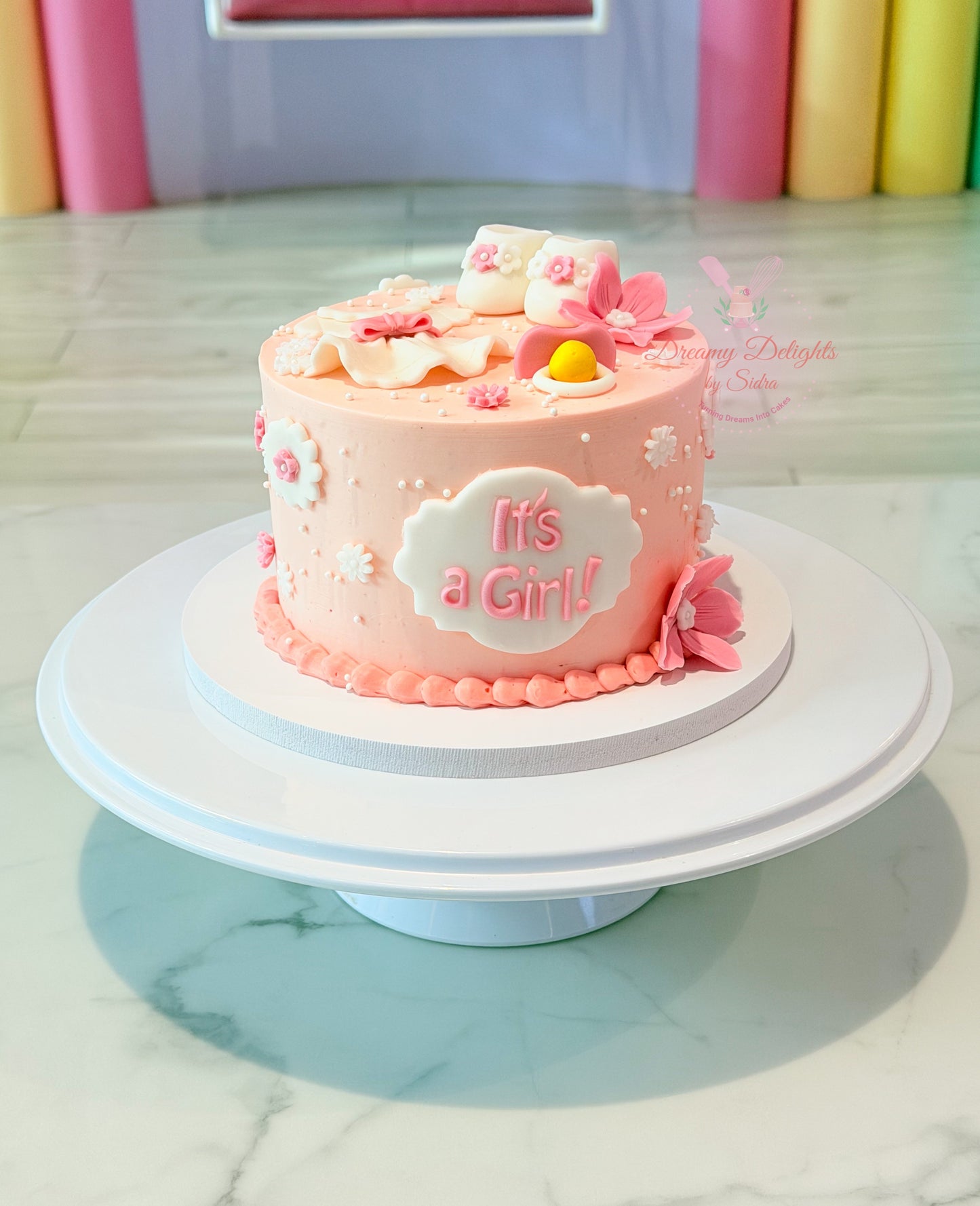 Pink It's a Girl baby shower cake with handcrafted baby shoes, baby dress, bonnet, flowers, and baby shower decorations in Dubai.