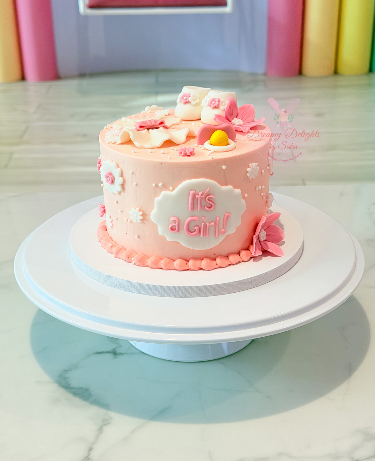 Pink It's a Girl baby shower cake with handcrafted baby shoes, baby dress, bonnet, flowers, and baby shower decorations in Dubai.