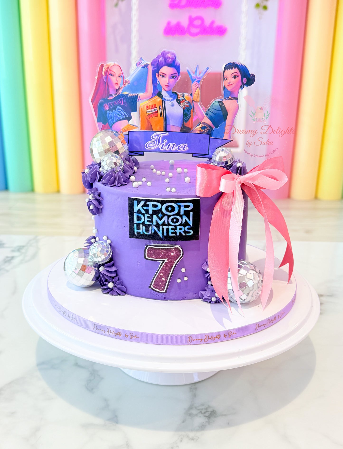 Personalized K-Pop ribbon birthday cake with purple buttercream, pink bow, disco ball decorations, and character toppers for girls in Dubai.