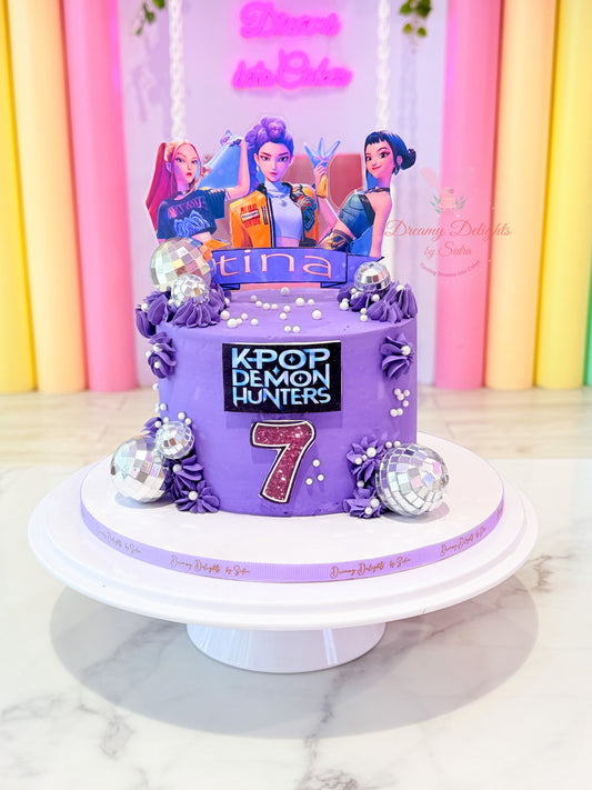 A purple K-pop themed birthday cake on a white cake stand, featuring character toppers, a 'tina' name sign, and a '7' decoration, set against a colorful pink and yellow curtain backdrop.