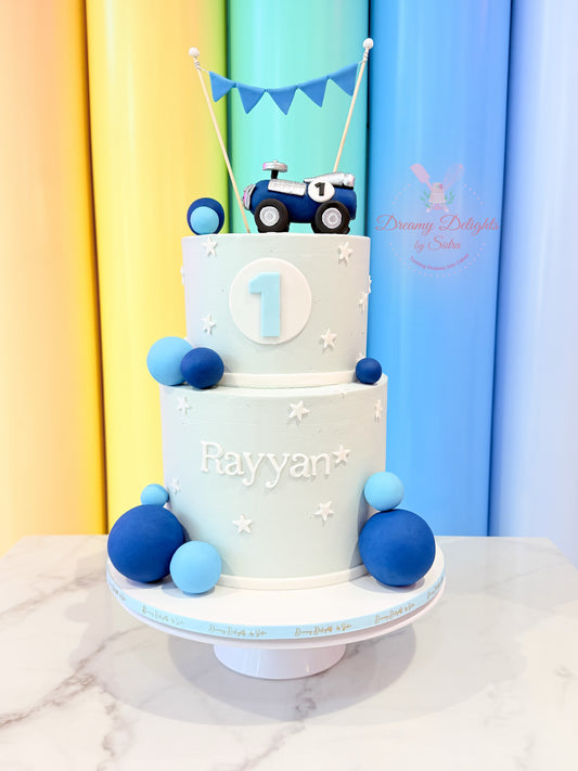 Little car first birthday cake with a blue car topper, white frosting, blue fondant balls, stars and personalised name by Dreamy Delights by Sidra in Dubai.