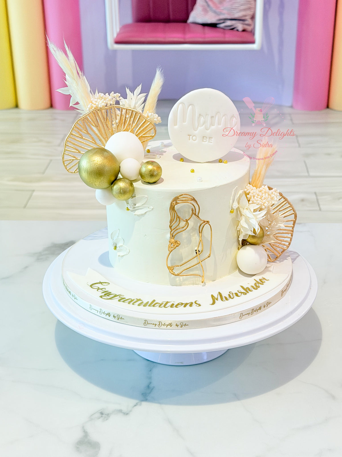 Elegant Mama to Be baby bump cake with white buttercream, gold accents, dried florals, and an acrylic maternity figurine by Dreamy Delights by Sidra in Dubai