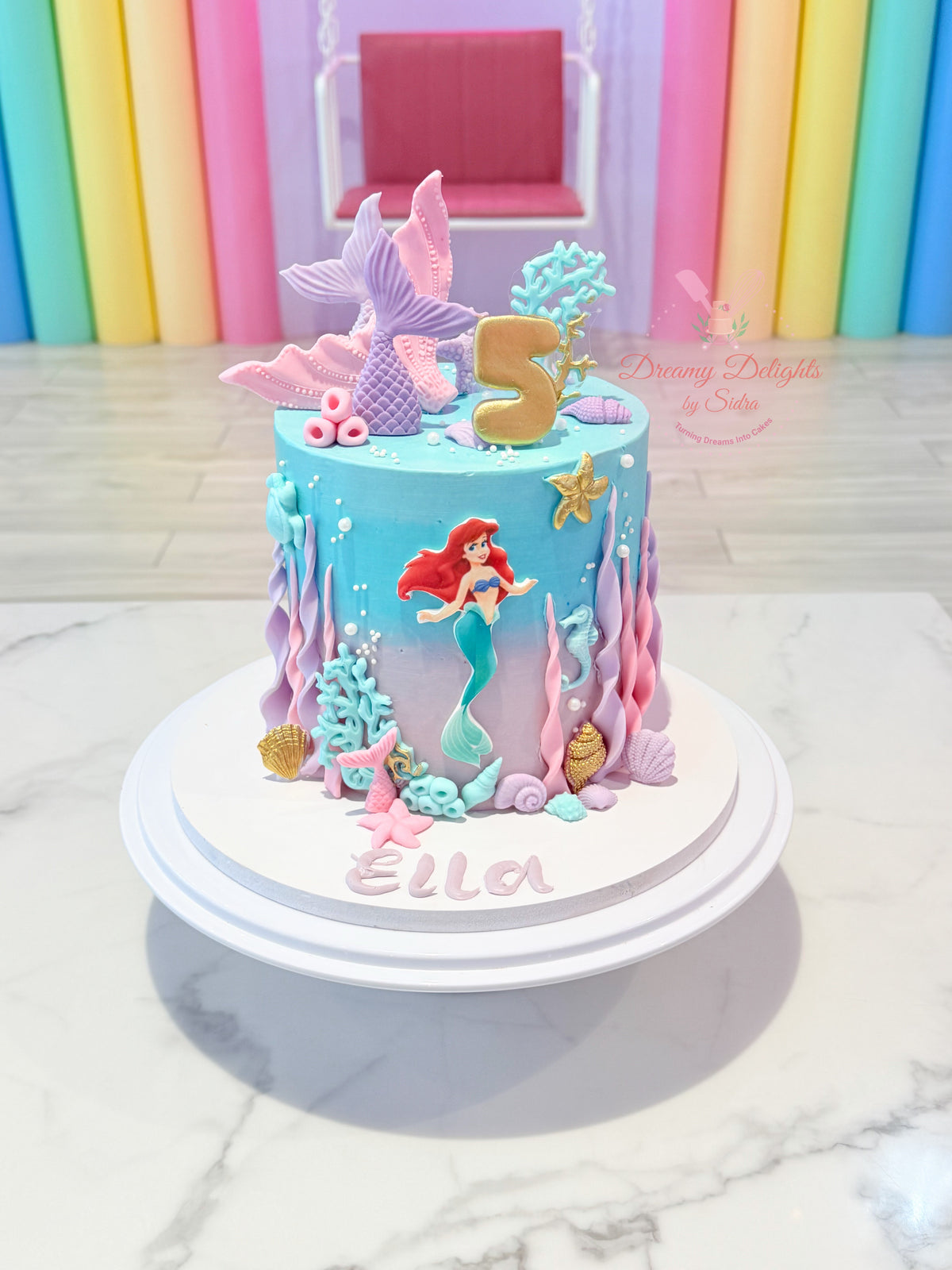 A custom mermaid-themed 5th birthday cake with Ariel decoration, a gold number 5 topper, and pink and purple sea-themed decorations, displayed on a white cake stand.