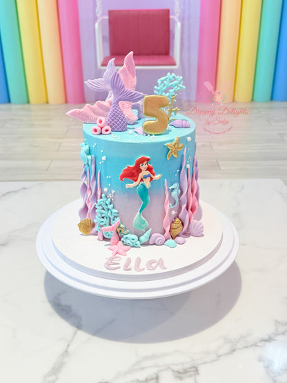A custom mermaid-themed 5th birthday cake with Ariel decoration, a gold number 5 topper, and pink and purple sea-themed decorations, displayed on a white cake stand.