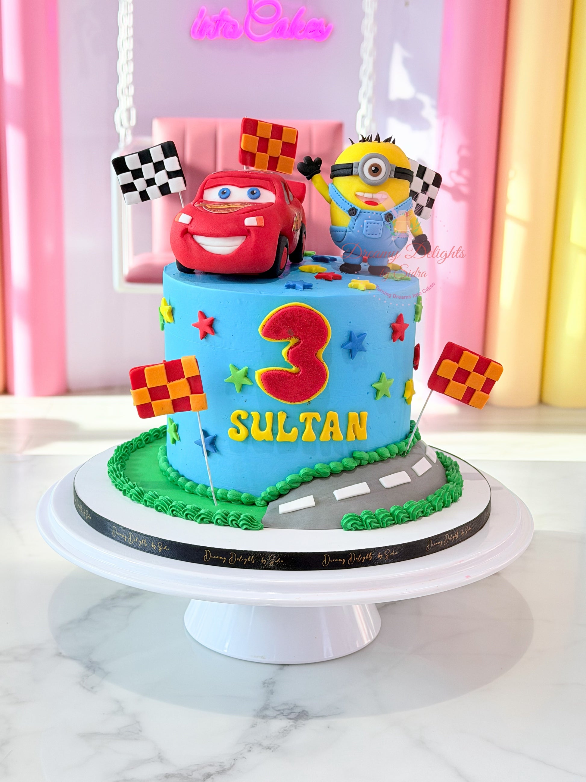 Personalized Minions and Lightning McQueen birthday cake with racing flags, colorful stars, road design, and custom name topper for boys in Dubai.
