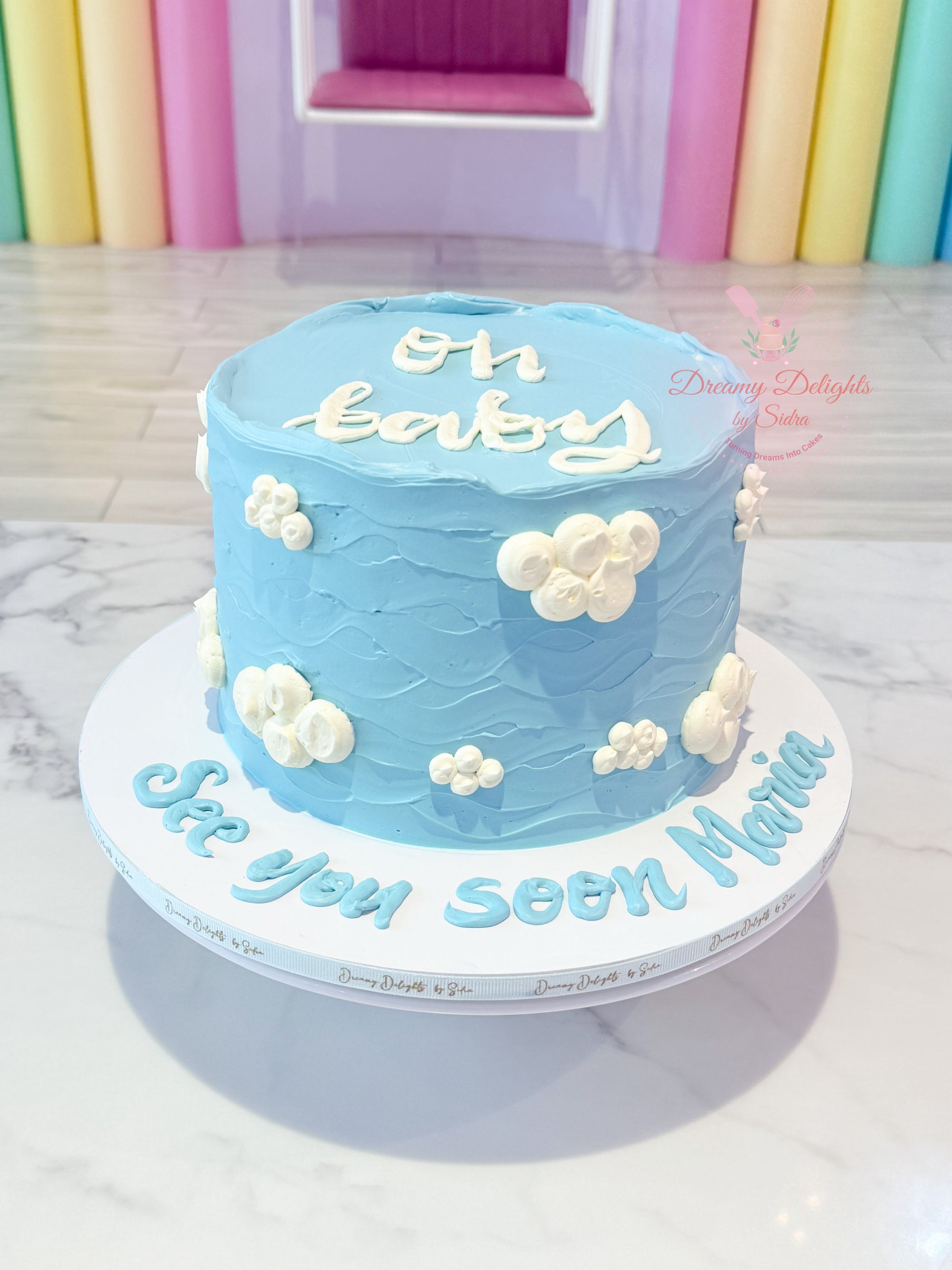 Blue baby shower cake with white text and clouds on a white stand.