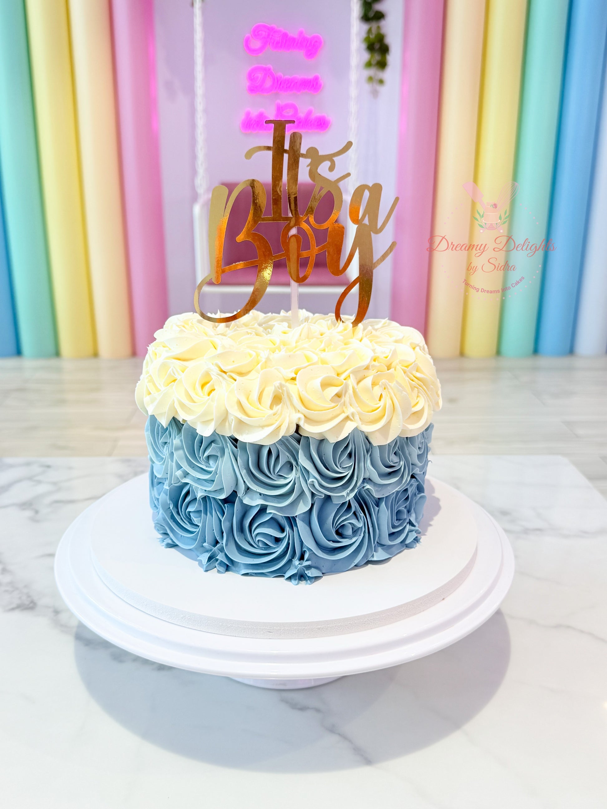 Two-tiered cake with rosette design and 'It's a Boy' topper against a pastel curtain background