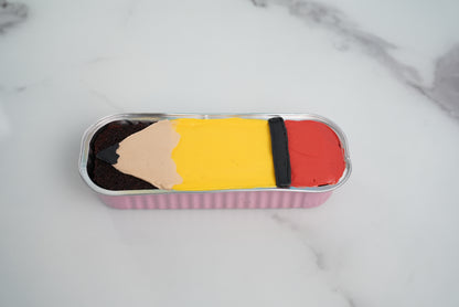A novelty pencil-shaped dessert in a pink foil container, featuring dark brown chocolate frosting, a beige frosting tip, a yellow frosting body, a black stripe, and a red frosting eraser, set on a white marble surface.
