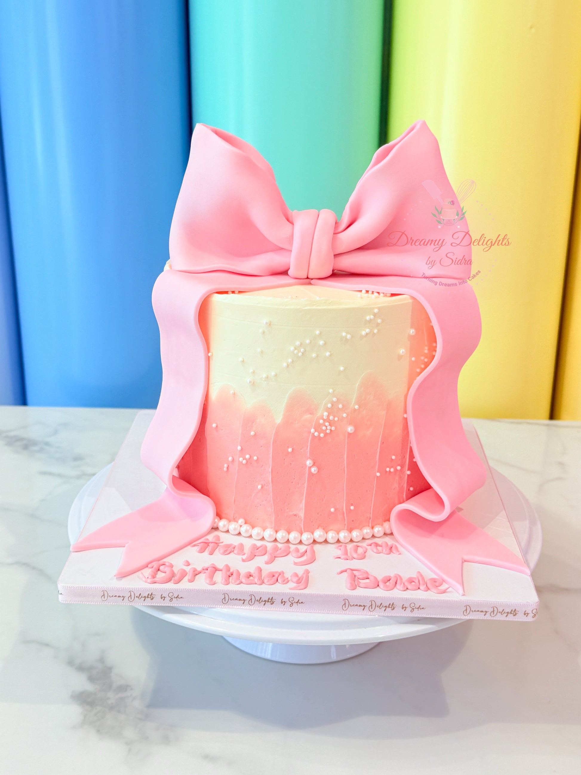 Pink birthday cake with a large bow and 'Happy Birthday' message against a colorful curtain background.