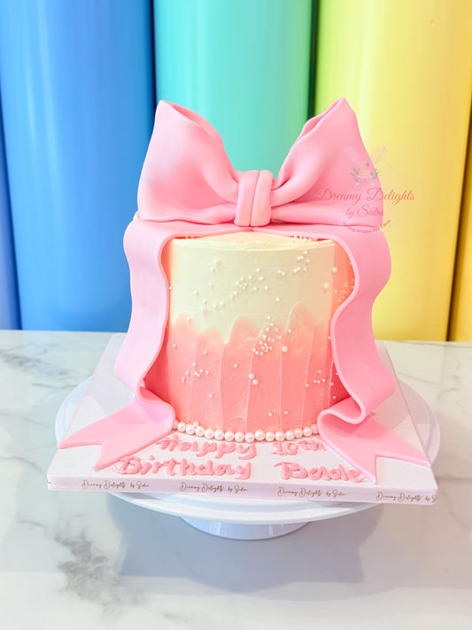 Pink birthday cake with a large bow and 'Happy Birthday' message against a colorful curtain background.