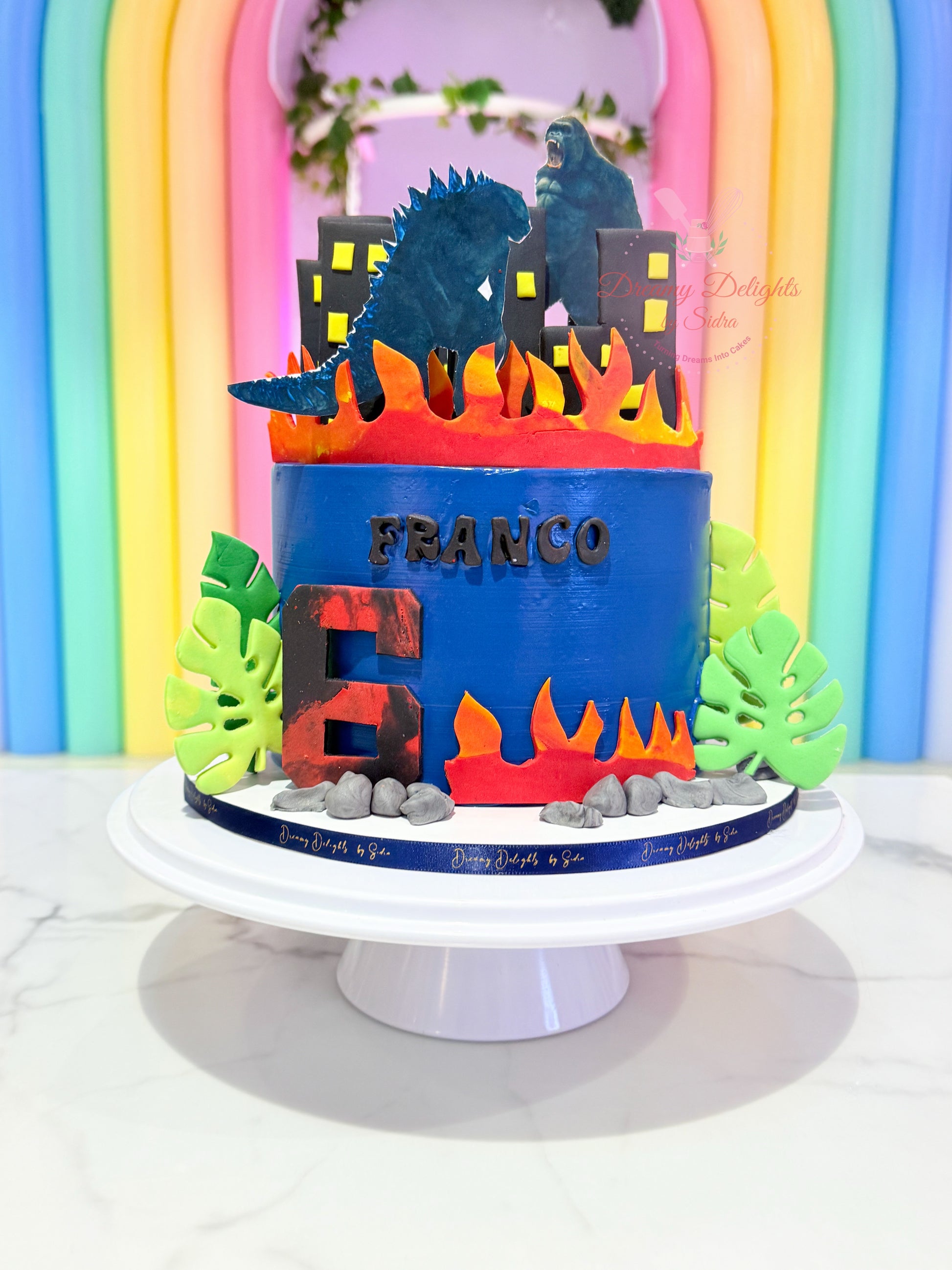 Colorful birthday cake with dinosaur design on a white stand against a rainbow-colored backdrop