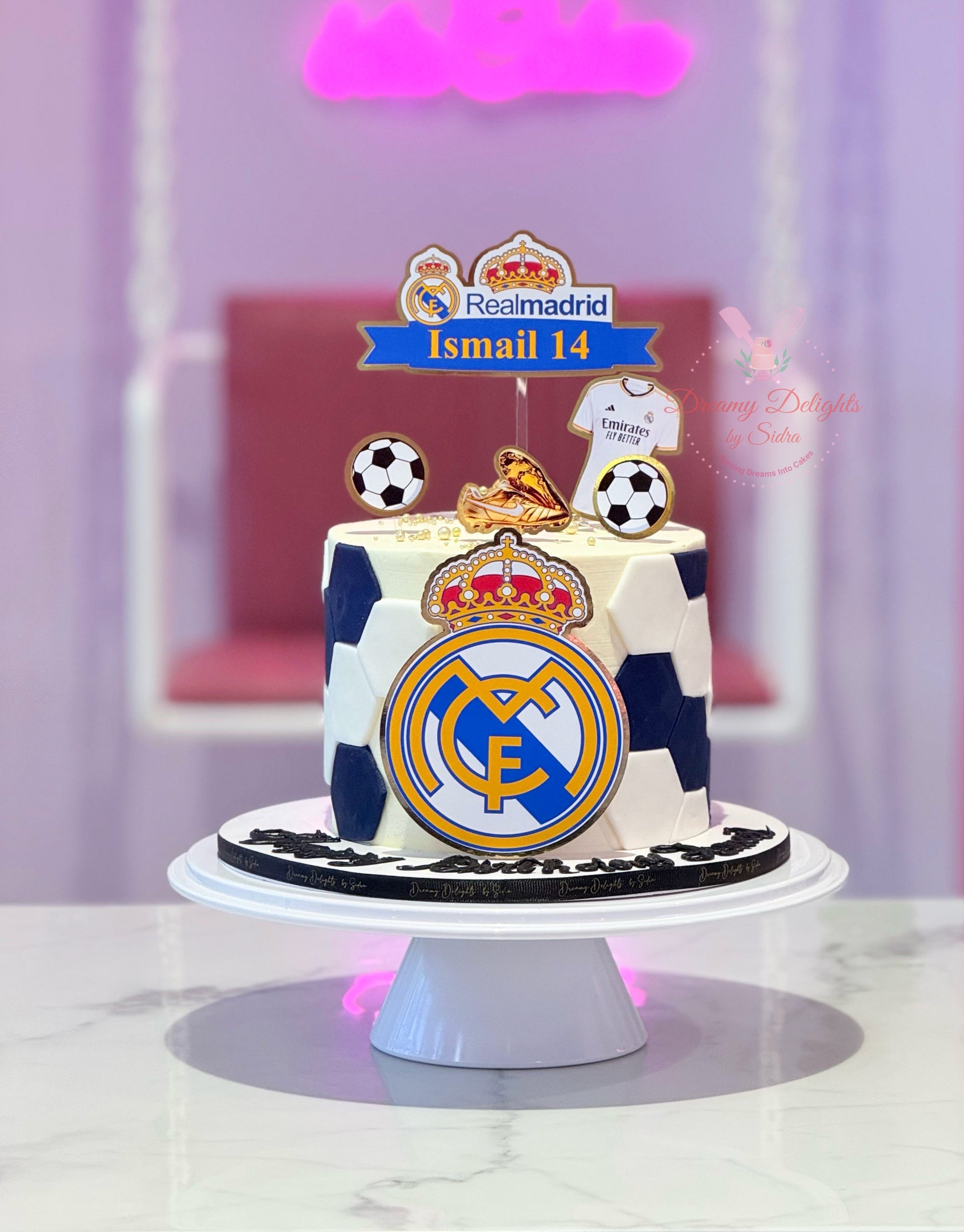 Cake with Real Madrid logo and soccer ball decorations on a white stand.