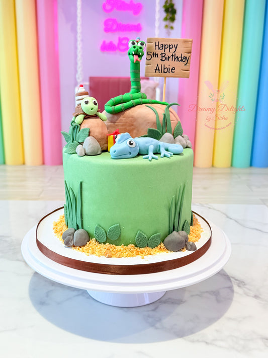 Children's birthday cake with green fondant, small figures, and 'Happy 5th Birthday Abbie' sign against a colorful curtain background.