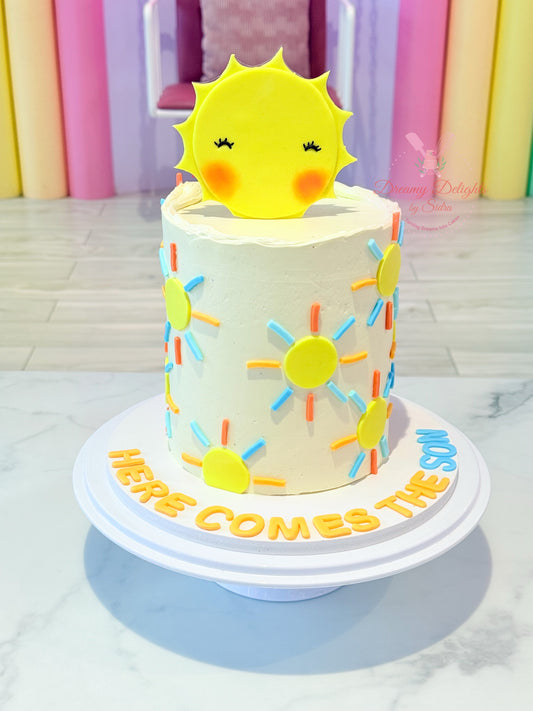 Decorative cake with sun design and 'Here Comes The Sun' text against a colorful curtain background