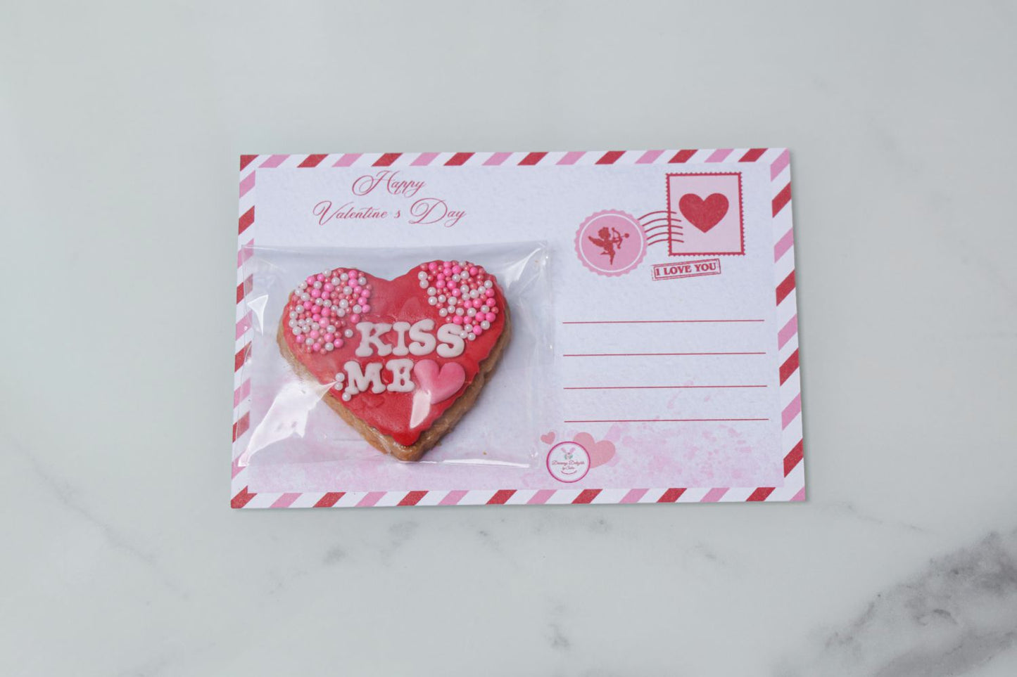 Valentine’s Day heart cookie with Kiss Me message attached to a postcard by Dreamy Delight by Sidra